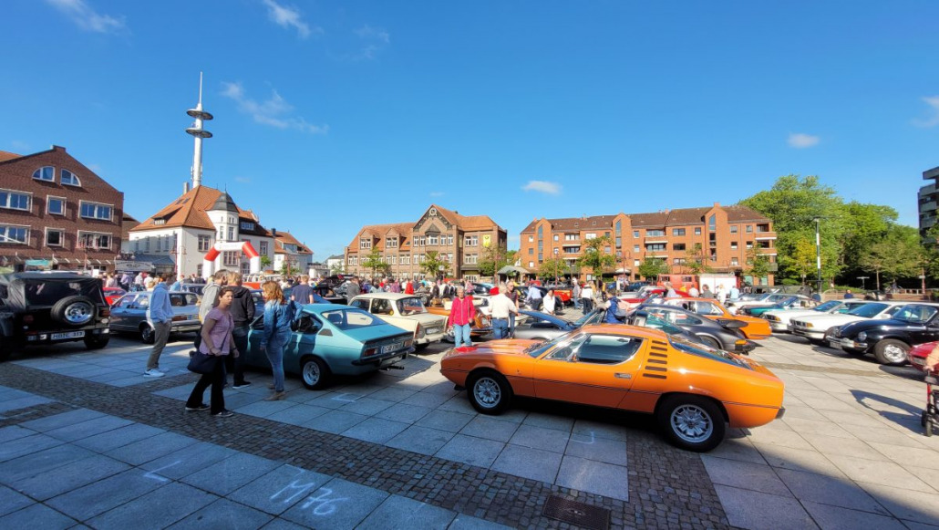 Eine Menschenmenge versammelt sich um Oldtimer, die auf einem sonnigen Platz im Freien mit umliegenden Gebäuden und einem hohen Turm im Hintergrund ausgestellt sind.